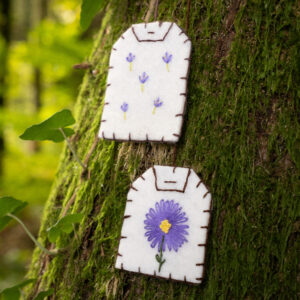 Gros plan sur deux marque-pages brodés en forme de sachet de thé (broderies aster violet et lavande) posés sur un tronc d'arbre moussu.
