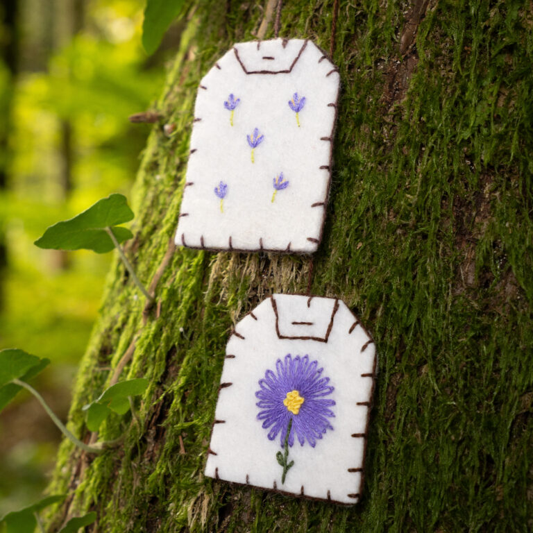 Gros plan sur deux marque-pages brodés en forme de sachet de thé (broderies aster violet et lavande) posés sur un tronc d'arbre moussu.