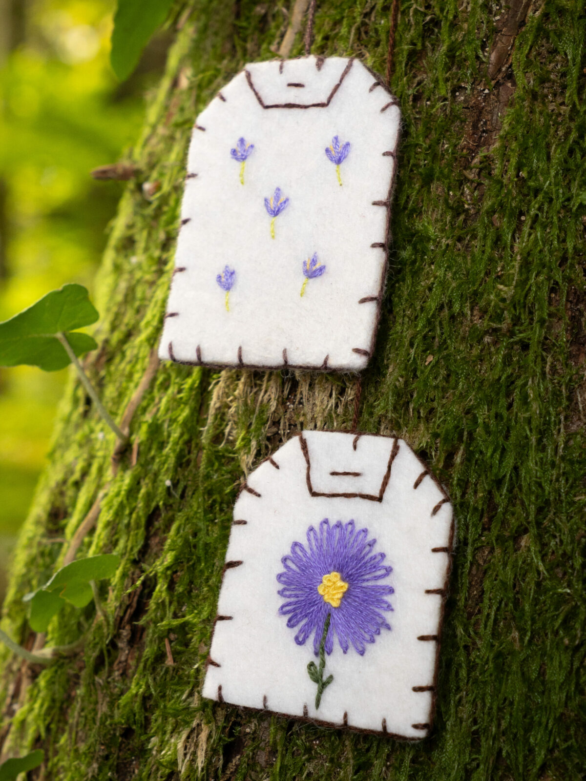 Gros plan sur deux marque-pages brodés en forme de sachet de thé (broderies aster violet et lavande) posés sur un tronc d'arbre moussu.