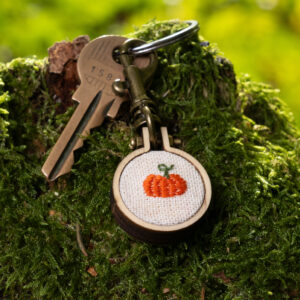Porte clé brodé citrouille en mini-tambour de bois, accroché à des clés sur un tronc d'arbre moussu.