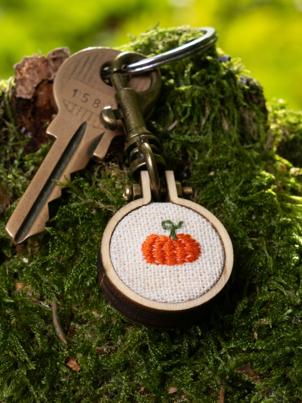 Porte clé brodé citrouille en mini-tambour de bois, accroché à des clés sur un tronc d'arbre moussu.